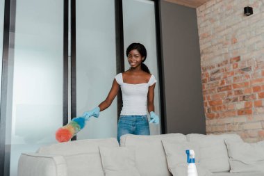 Plastik eldivenli Afrikalı Amerikalı kadın oturma odasını toz beziyle temizliyor.