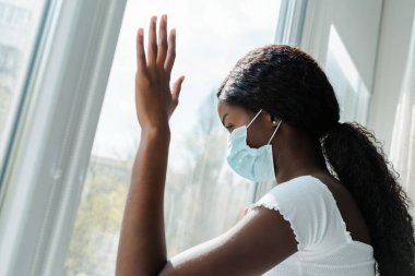 Tıbbi maskeli Afrikalı Amerikalı kız oturma odasındaki pencerelere dokunuyor.