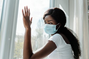 Tıbbi maskeli Afrikalı Amerikalı kız pencerelere dokunuyor ve oturma odasındaki kameraya bakıyor.