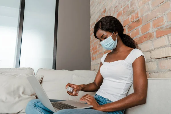 医療用マスクのアフリカ系アメリカ人のフリーランスは リビングルームでソファの上にハンドサニタイザーでノートパソコンを消毒 — ストック写真