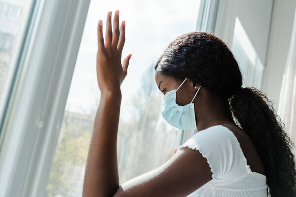 African american girl in medical mask touching windows in living room