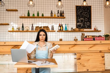 Kafe sahibi kameraya bakıyor ve dizüstü bilgisayarın yanında kağıt tutuyor ve masada gözlüklü belgeler var.