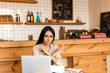 Cafe owner looking at camera and showing no sign near laptop, calculator and papers at table clipart