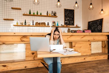 Stressed cafe owner looking at laptop near documents at table clipart