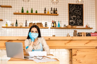 Masadaki gözlük, kağıt ve dizüstü bilgisayara bakan tıbbi maskeli bir kafe sahibi.