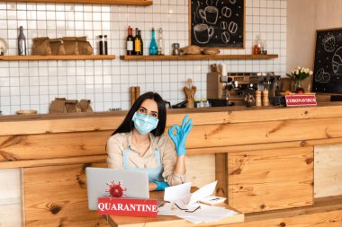 Tıbbi maskeli bir kafe sahibi kameraya bakıyor ve masada karantina harfli kağıtlar, dizüstü bilgisayarlar, bardaklar ve kartların yanında onay işareti gösteriyor.