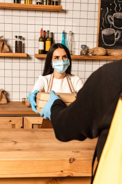 Medikal maskeli kafe sahibinin seçici odak noktası masa yakınındaki kuryeye kutu vermek