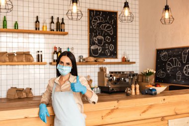 Tıbbi maskeli kafe sahibi ve lateks eldivenli. Kameraya bakıyor ve tabela gibi bir şey gösteriyor.