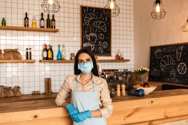 Maskenin yanındaki kameraya bakan tıbbi maskeli ve lateks eldivenli kafe sahibi.