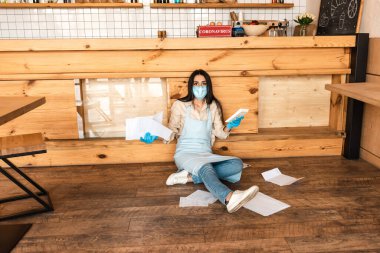 Medikal maskeli bir kafe sahibi hesap makinesiyle dokümanları tutuyor ve masanın yanındaki kameraya bakıyor. 