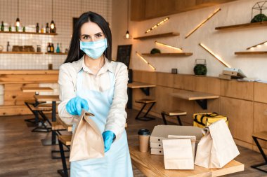 Tıbbi maskeli bir kafe sahibi kağıt poşet gösteriyor ve masanın yanındaki tek kullanımlık kahve ve kutularla kameraya bakıyor.