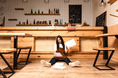Concentrated cafe owner looking at document and sitting near table on floor clipart