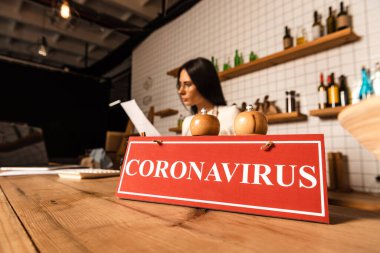 Selective focus of cafe owner looking at paper near card with coronavirus lettering with salt and pepper mills on table clipart