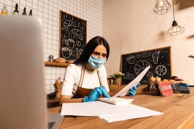 Masanın yanında hesap makinesi kullanan tıbbi maskeli endişeli bir kafe sahibi. 