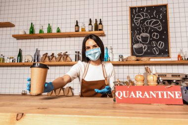 Tıbbi maskeli bir kafe sahibi kameraya bakıyor ve karantina yazılı bir kartla masanın yanındaki kahve bardağını işaret ediyor.