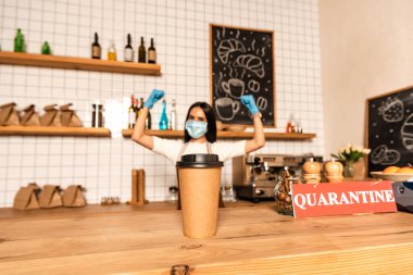 Selective focus of paper cup of coffee near card with quarantine lettering on table and cafe owner in medical mask with hands in air clipart