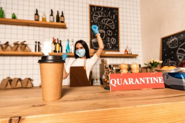 Selective focus of disposable cup of coffee near card with quarantine lettering on table and cafe owner in medical mask with hand in air clipart