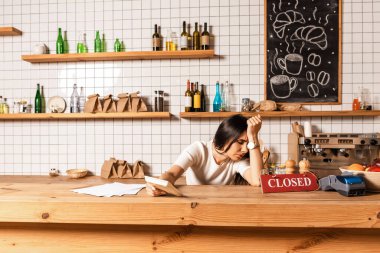 Upset cafe owner with closed eyes holding calculator and leaning on table with papers, card with closed lettering and payment terminal clipart