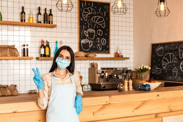 Cafe owner in medical mask and latex gloves looking at camera and showing victory sign near table