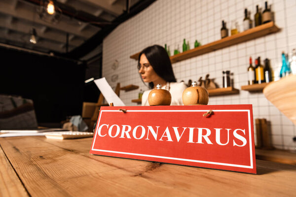 Selective focus of cafe owner looking at paper near card with coronavirus lettering with salt and pepper mills on table