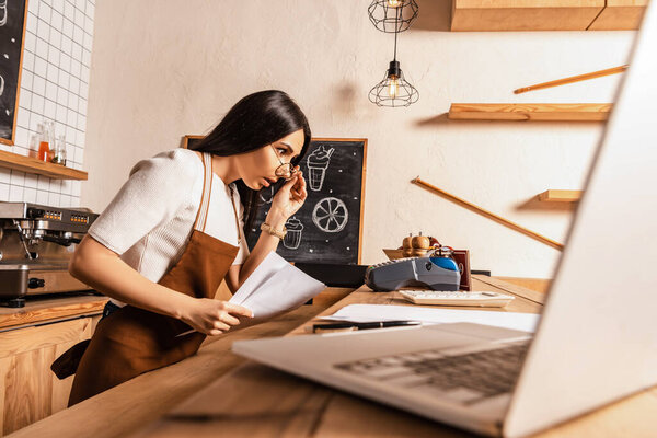 Selective focus of shocked cafe owner with papers looking at calculator near payment terminal and laptop on table