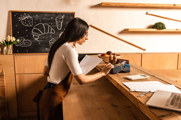 Side view of concentrated cafe owner with papers using payment terminal near calculator on table