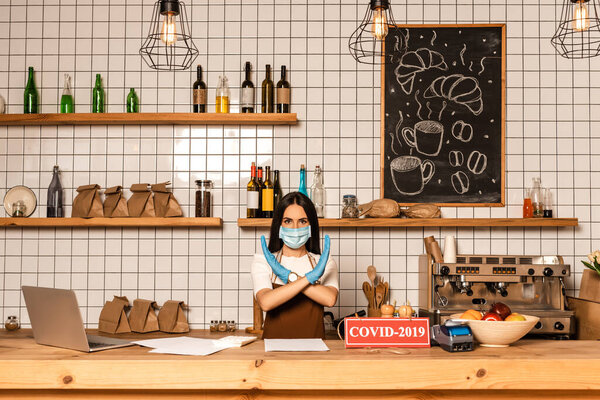 Cafe owner in medical mask showing no sign near table with papers, card with covid-2019 lettering, payment terminal and bowl with fruits