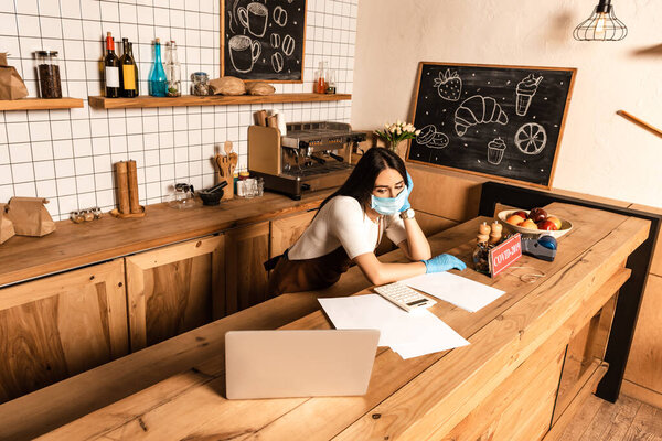 Высокоугольный вид владельца кафе рядом со столом с ноутбуком, бумагами, картой, платежным терминалом и чашей с фруктами
 