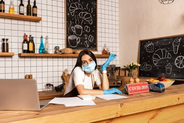 Kafe sahibi masanın yanındaki masaya dizüstü bilgisayarlı, kağıtlı, kartlı, covid-2019 harfli ödeme terminali ve meyve tabağıyla bakıyor. 