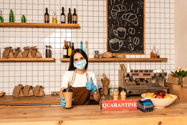 Cafe owner in medical mask showing like sign and paper cup of coffee near table with payment terminal, card with quarantine lettering and bowl with fruits