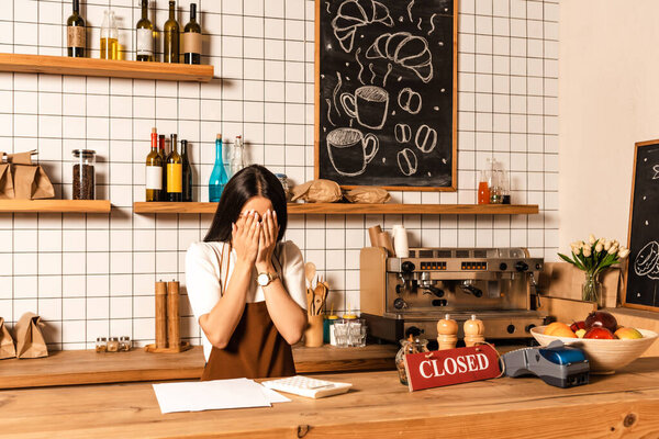 Cafe owner covering face near table with papers, calculator, card with closed lettering, payment terminal and bowl with fruits 