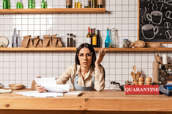 Worried cafe owner holding papers near calculator and card with quarantine lettering and looking at camera near table