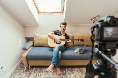 Genç video blogcusu dizüstü bilgisayarın yanındaki koltukta oturup dijital kameraya bakarken gitar çalıyor.