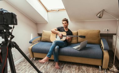 Ciddi video blogcusu dizüstü bilgisayarın yanındaki koltukta oturup dijital kameraya bakarken gitar çalıyor.