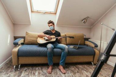 Koruyucu maskeli genç adam tavan arası odasındaki dizüstü bilgisayarın yanında otururken gitar çalıyor.