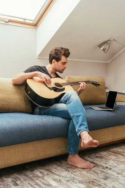 Atento Joven Tocando Guitarra Mientras Está Sentado Sofá Cerca Computadora —  Fotos de Stock