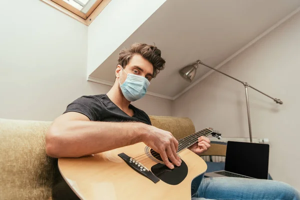 医療用マスクの若者がギターを弾きながらカメラを見ながらノートパソコンのそばのソファに空白の画面で座っています — ストック写真