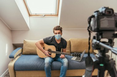 Tıbbi maskeli genç bir vlogger 'ın tripoddaki dijital kameraya bakarken gitar çalmasının seçici odağı