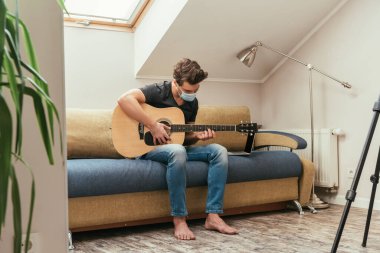 Maskeli genç adam dizüstü bilgisayarın yanındaki koltukta otururken gitar çalıyor.