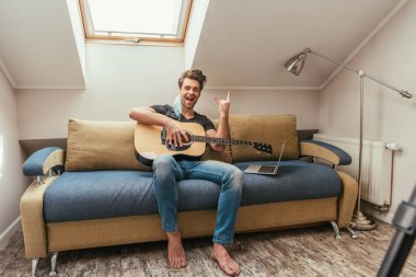 Kulağında gitar tutan ve dizüstü bilgisayarın yanındaki koltukta otururken kaya işareti gösteren tıbbi maskeli heyecanlı bir adam. 