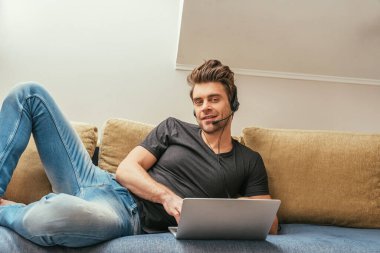 Kulaklıklı yakışıklı adam dizüstü bilgisayarın yanındaki koltukta uzanıyor ve kameraya gülümsüyor.