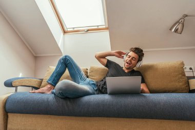 Dizüstü bilgisayarın yanındaki kanepede yatarken kablosuz kulaklıkla müzik dinleyen heyecanlı bir adam.