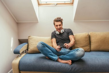 KYIV, UKRAINE - 13 Nisan 2019: bacak bacak bacak üstüne atmış kanepede otururken boynunda kablosuz kulaklık olan heyecanlı adam video oyunu oynuyor.