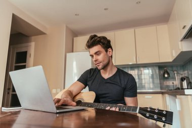 Gitarlı yakışıklı genç adam mutfakta dizüstü bilgisayar kullanıyor.