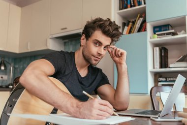 Gitar çalan yakışıklı genç adam dizüstü bilgisayarın yanında otururken kameraya bakıyor.