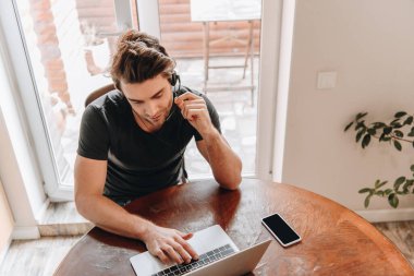 akıllı telefonun yanında dizüstü bilgisayar kullanan kulaklıklı genç adamın yüksek açılı görüntüsü ve evde boş ekran var.