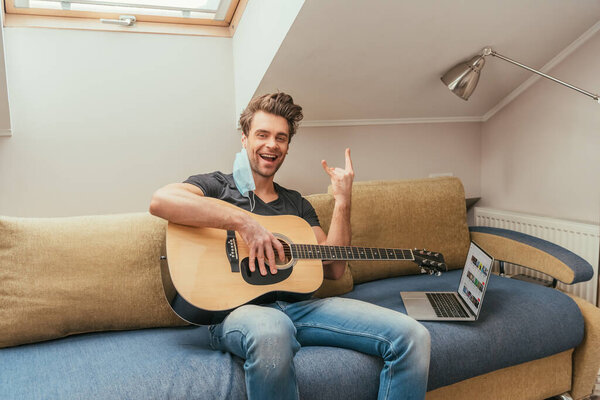 KYIV, UKRAINE - APRIL 13, 2019: cheerful man with protective mask on ear holding guitar and showing rock sign while sitting on sofa near laptop with Youtube on screen