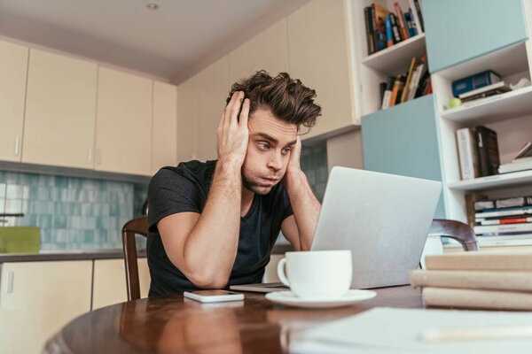 exhausted man touching head while looking at laptop monitor near cup of coffee