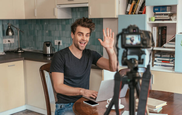 selective focus of young vlogger waving hand in kitchen while looking at digital camera on tripod