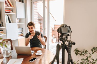 Bilgisayarın yanında otururken parmaklarıyla dijital kamerayı gösteren neşeli vlogger